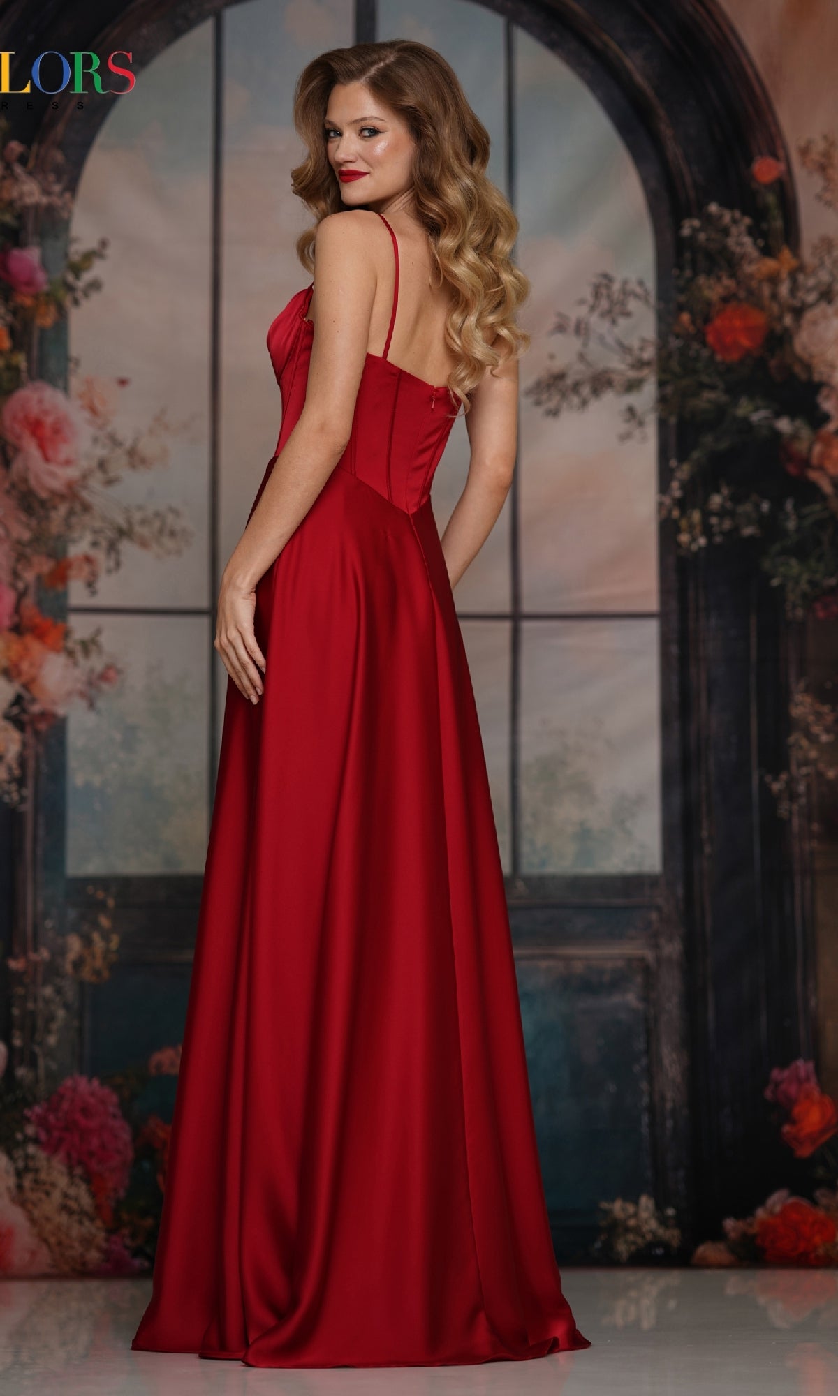 A woman with wavy hair models the Long Satin Corset Prom Dress: Colors Dress 3743, standing before a floral arched window backdrop and glancing over her shoulder.